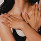 A close-up lifestyle shot of a woman wearing multiple gold rings on her hands and neck, posing against a neutral light gray background.