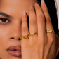 A close-up of a woman with dark hair wearing gold rings on her fingers, holding her hand near her face against a plain light background.