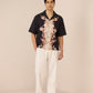Man wearing a black floral shirt and white pants on a beige background Calling June