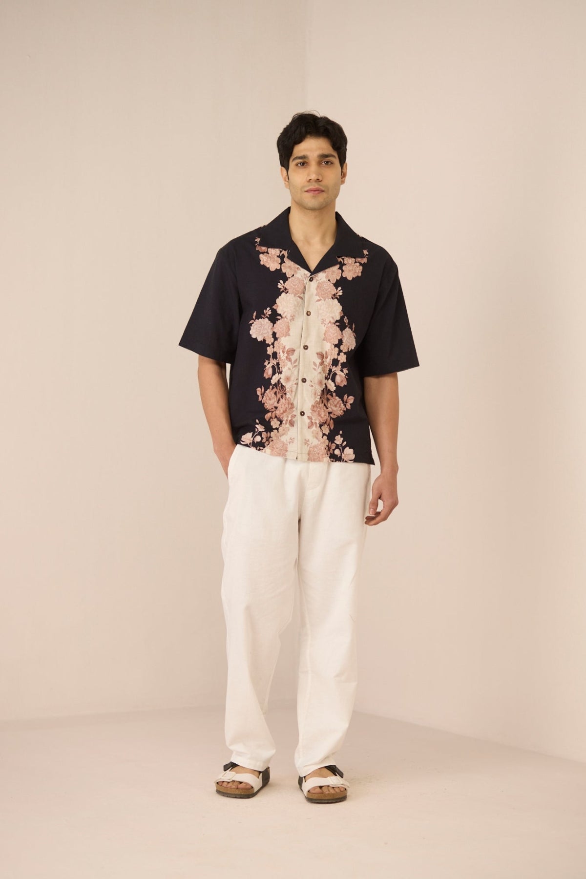 Man wearing a black floral shirt and white pants on a beige background Calling June