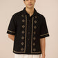 Man wearing a black embroidered shirt with white pants on a beige background Calling June