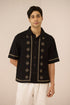 Man wearing a black embroidered shirt with white pants on a beige background Calling June
