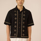 Man wearing a black embroidered shirt with white pants on a beige background Calling June