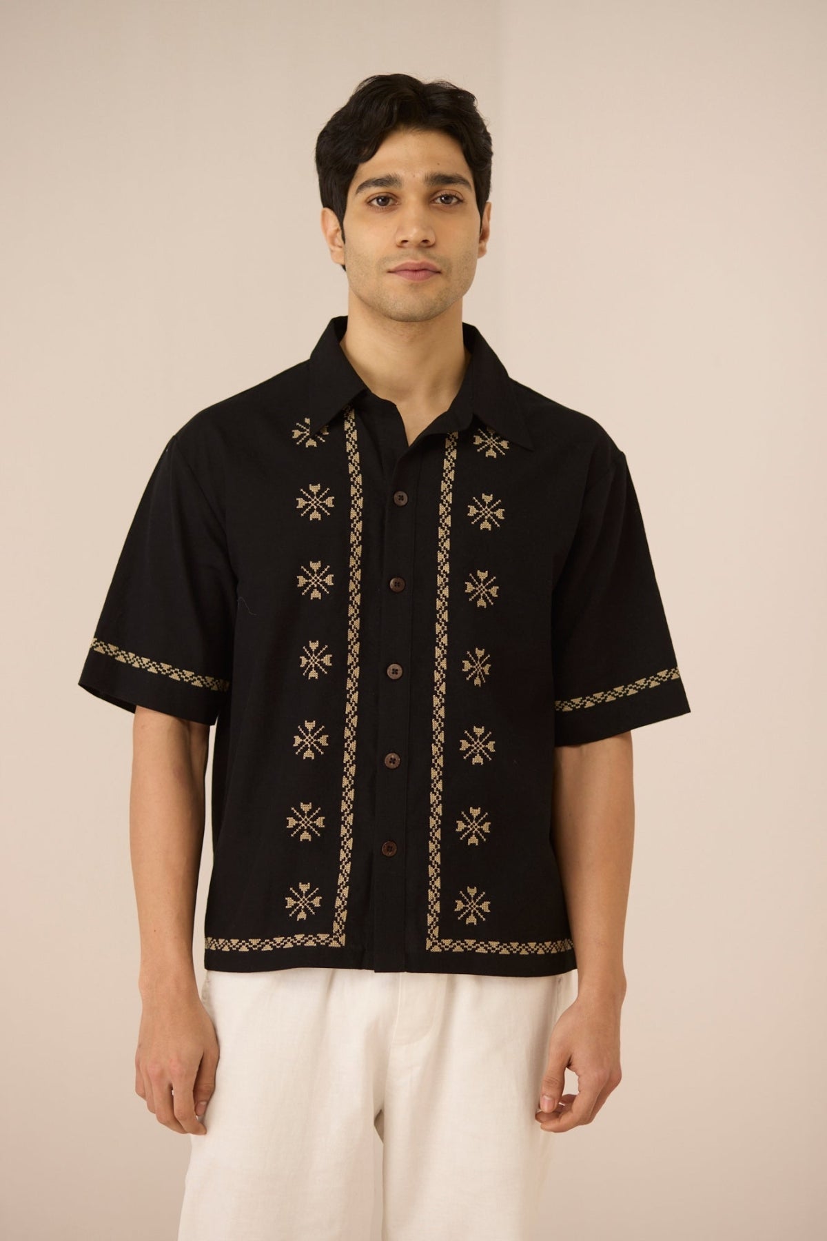 Man wearing a black embroidered shirt with white pants on a beige background Calling June