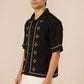 Man wearing a black embroidered shirt with white pants on a beige background Calling June