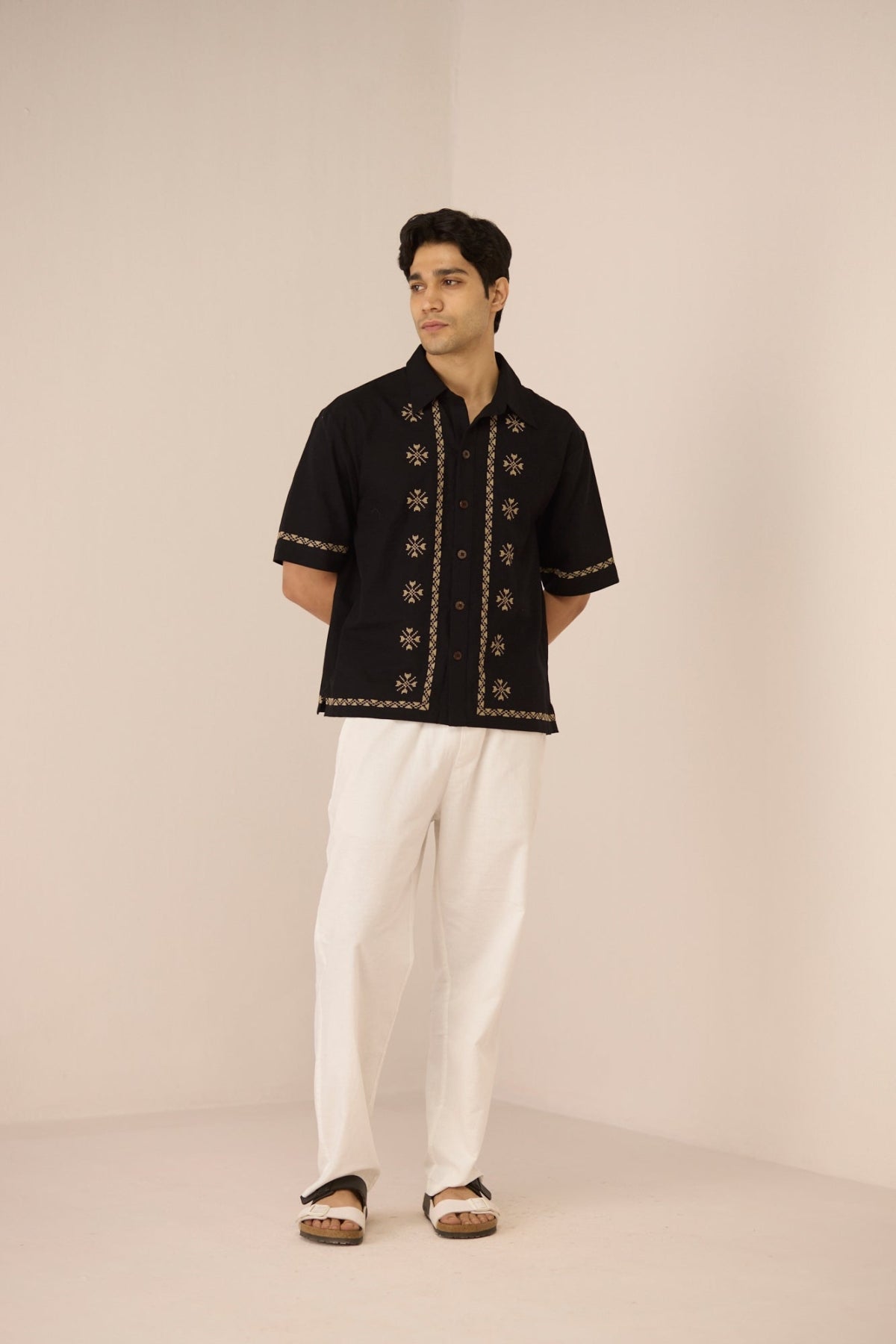 Man wearing a black embroidered shirt and white pants on a beige background Calling June