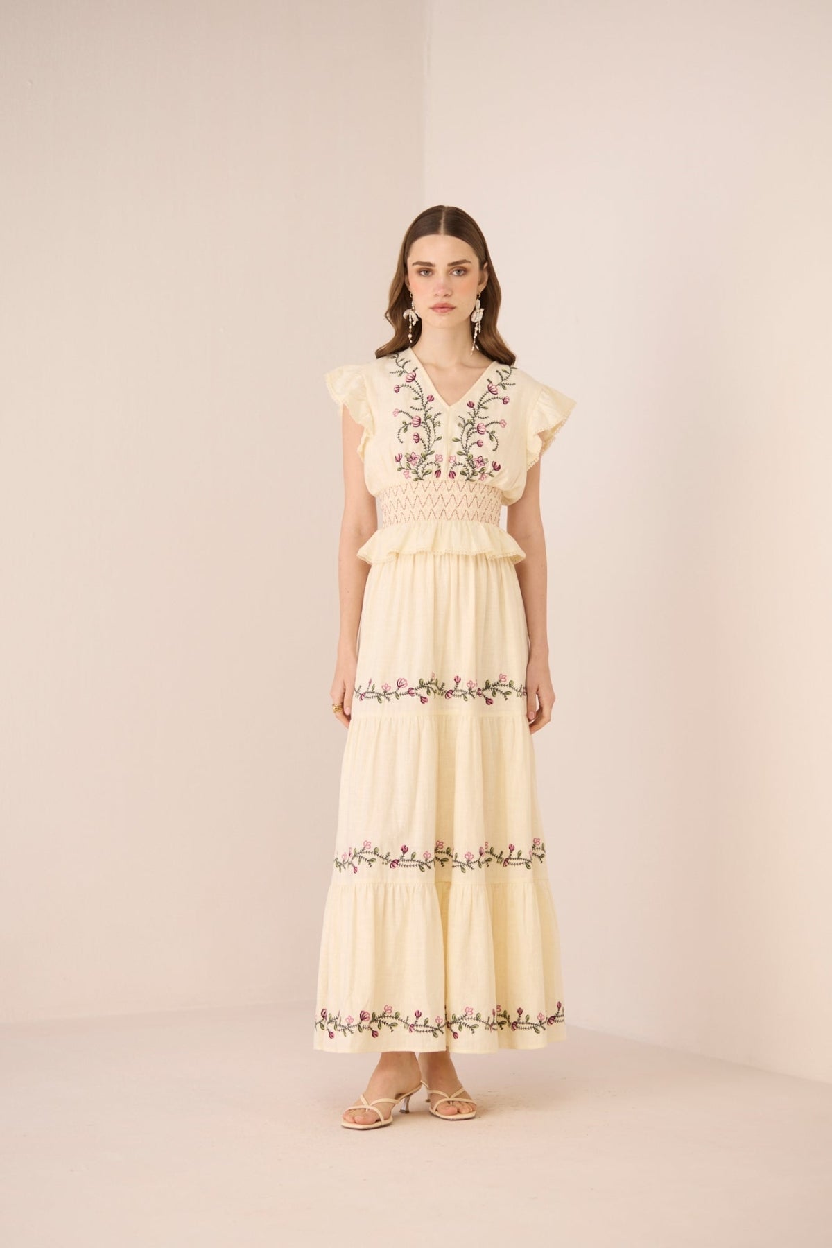 Woman wearing a long, light-colored dress with floral embroidery on a plain background Calling June