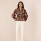Woman wearing a patterned blouse and white pants against a plain background Calling June