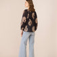 Woman wearing a black blouse with gold patterns and light blue jeans on a beige background Calling June