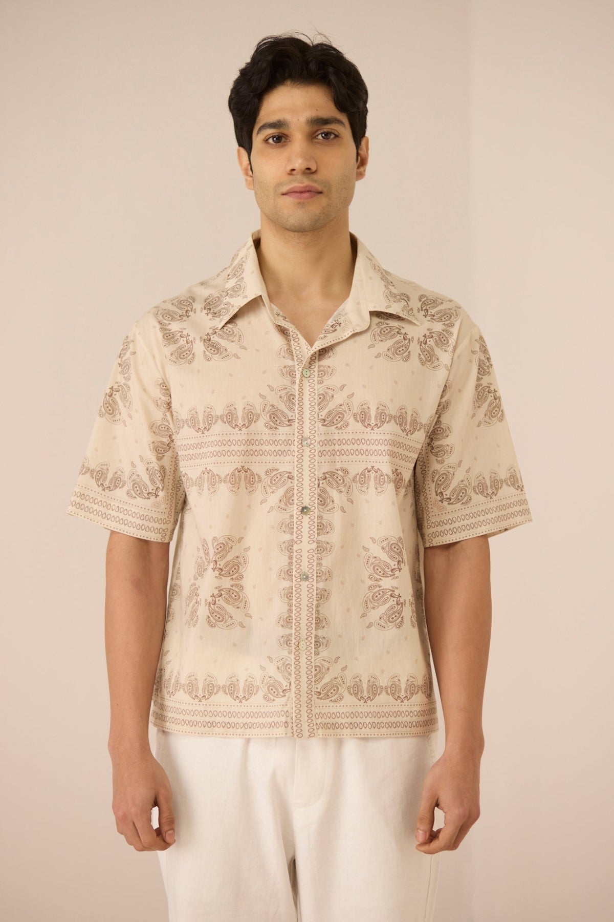 Man wearing a beige patterned shirt and white pants against a plain background Calling June