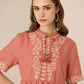 Woman wearing a pink dress with white floral embroidery against a beige background Calling June