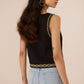 Woman wearing a black sleeveless top with gold detailing and light blue jeans on a beige background Calling June