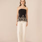 Woman wearing a black strapless top with white embroidery and white pants on a beige background Calling June