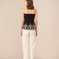 Woman wearing a black strapless top with lace detailing and white pants on a beige background Calling June