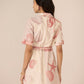 Woman wearing a light-colored dress with pink floral patterns on a plain background Calling June