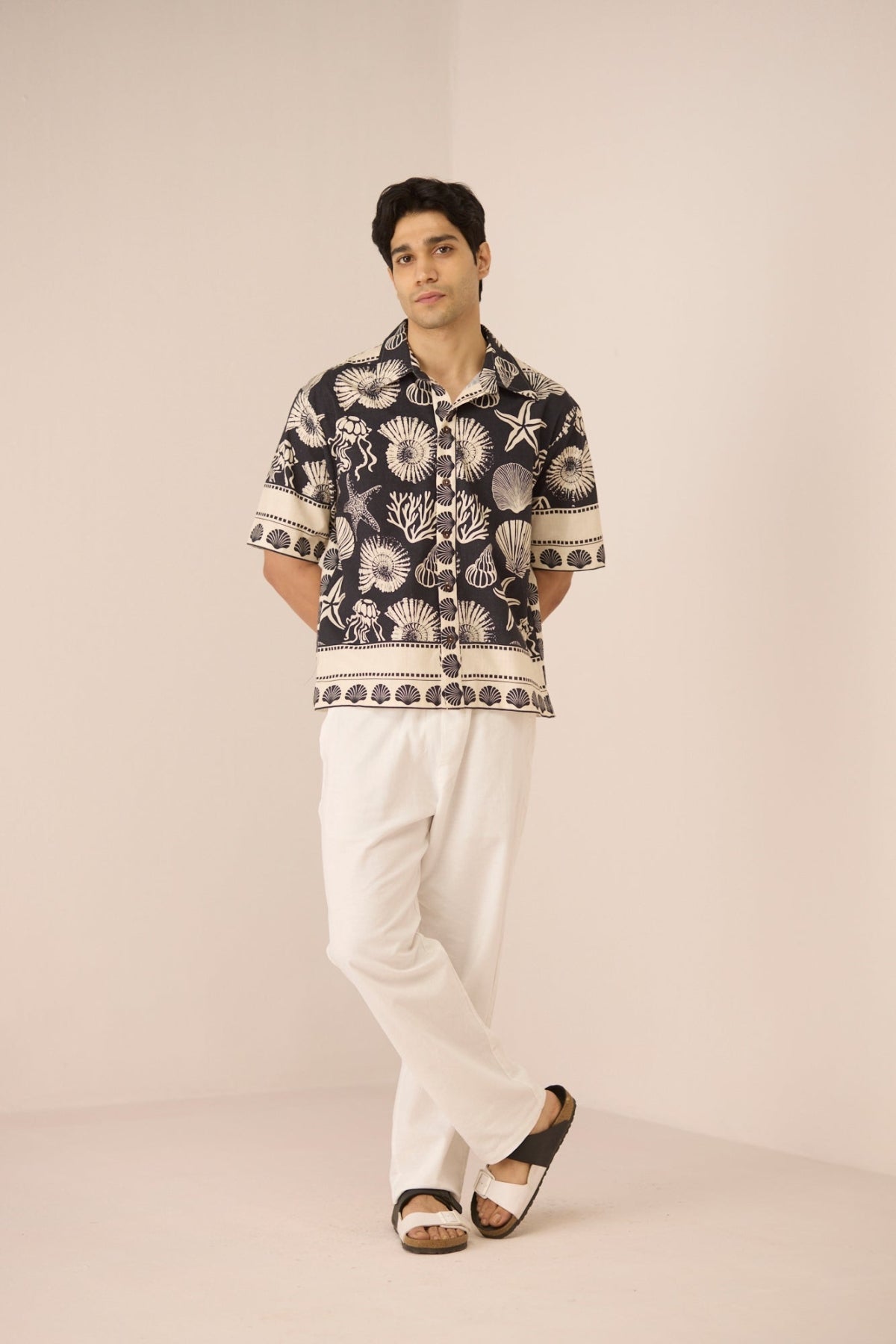 Man wearing a patterned shirt and white pants on a plain background Calling June