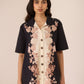 Woman wearing a black floral jacket over a cream shirt with matching pants on a beige background Calling June