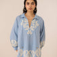Woman wearing a light blue blouse with intricate white embroidery on a beige background Calling June