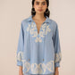 Woman wearing a light blue embroidered blouse against a beige background Calling June