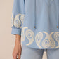 Light blue blouse with intricate white embroidery on a beige background Calling June