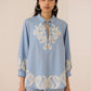 Woman wearing a light blue shirt with intricate white embroidery on a beige background Calling June
