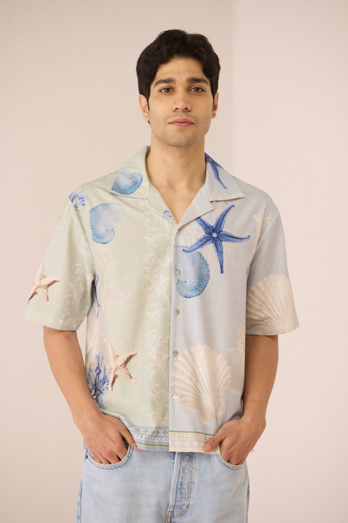 Man wearing a light-colored shirt with starfish and shell patterns on a plain background Calling June