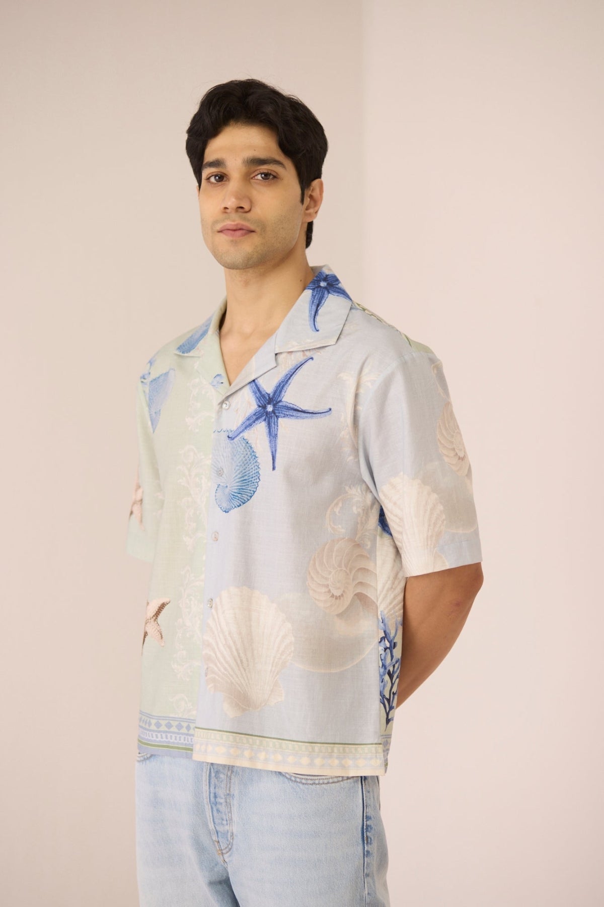 Man wearing a patterned shirt with starfish and shell designs on a plain background Calling June
