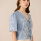 Woman wearing a light blue embroidered blouse against a beige background Calling June