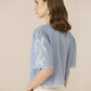 Woman wearing a light blue blouse with white embroidery on a beige background Calling June