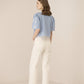 Woman wearing a light blue top with lace sleeves and white pants on a beige background Calling June